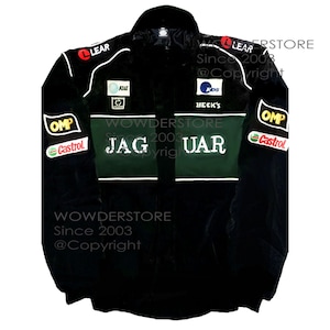 Op de afbeelding: Zwarte racejas met het woord "JAGUAR" in witte letters over de borst. De jas heeft groene en witte accenten, evenals sponsorlogo's zoals "OMP", "Castrol" en "LEAR". Het is een model met volledige ritssluiting en een opstaande kraag.