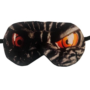 May include: A black sleep mask featuring a close-up of a monster's face with orange and red eyes. The mask has a black elastic band for securing it around the head. The design is printed on a soft fabric.