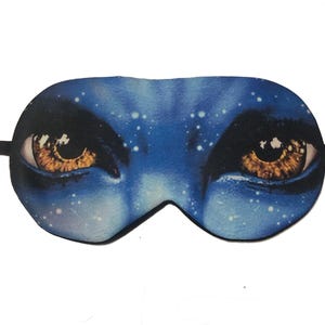 May include: A blue sleep mask featuring a detailed graphic of eyes and facial features. The eyes are a warm brown color, and the skin has a blue hue with white speckles. The mask has a black elastic band.