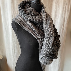 May include: A grey and white chunky knit scarf is displayed on a black mannequin. The scarf has a textured, open weave design. The neutral colour palette makes it a versatile accessory. The scarf is likely made of wool or a wool blend.