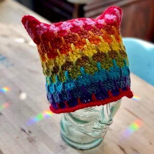 May include: A colorful, handmade crochet hat with cat ears. The hat features a rainbow of colors, including red, orange, yellow, green, blue, and purple. The hat is displayed on a glass head and has a red brim.