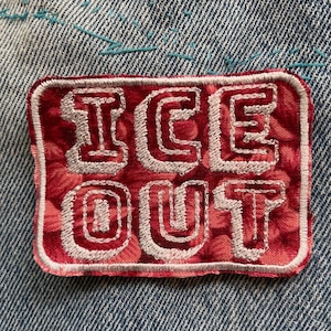 Puede incluir: Un parche de tela rectangular con un patrón moteado en rojo y blanco que presenta el texto blanco bordado ICE OUT en una fuente de bloque en negrita. El parche tiene un borde cosido en blanco y está sujeto a una base de tela vaquera azul.