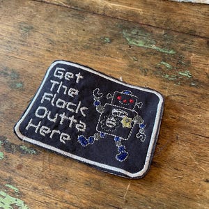 Puede incluir: Un parche rectangular negro bordado con un borde blanco y el texto blanco Get The Flack Outta Here. A la derecha del texto hay un pequeño personaje robot con ojos rojos, extremidades azules y un detalle amarillo en el pecho.
