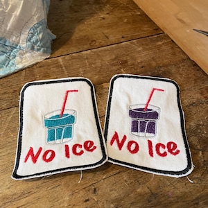 Può includere: Due toppe rettangolari bianche ricamate con bordi neri. Ogni toppa presenta un bicchiere con una cannuccia rossa e la scritta "No Ice" ricamata in rosso. Un bicchiere contiene liquido blu, l'altro contiene liquido viola.