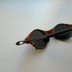 May include: Round sunglasses with dark lenses and a brown leather strap. The frame is dark, and the leather strap is stitched to the frame. The sunglasses have a unique, vintage style.