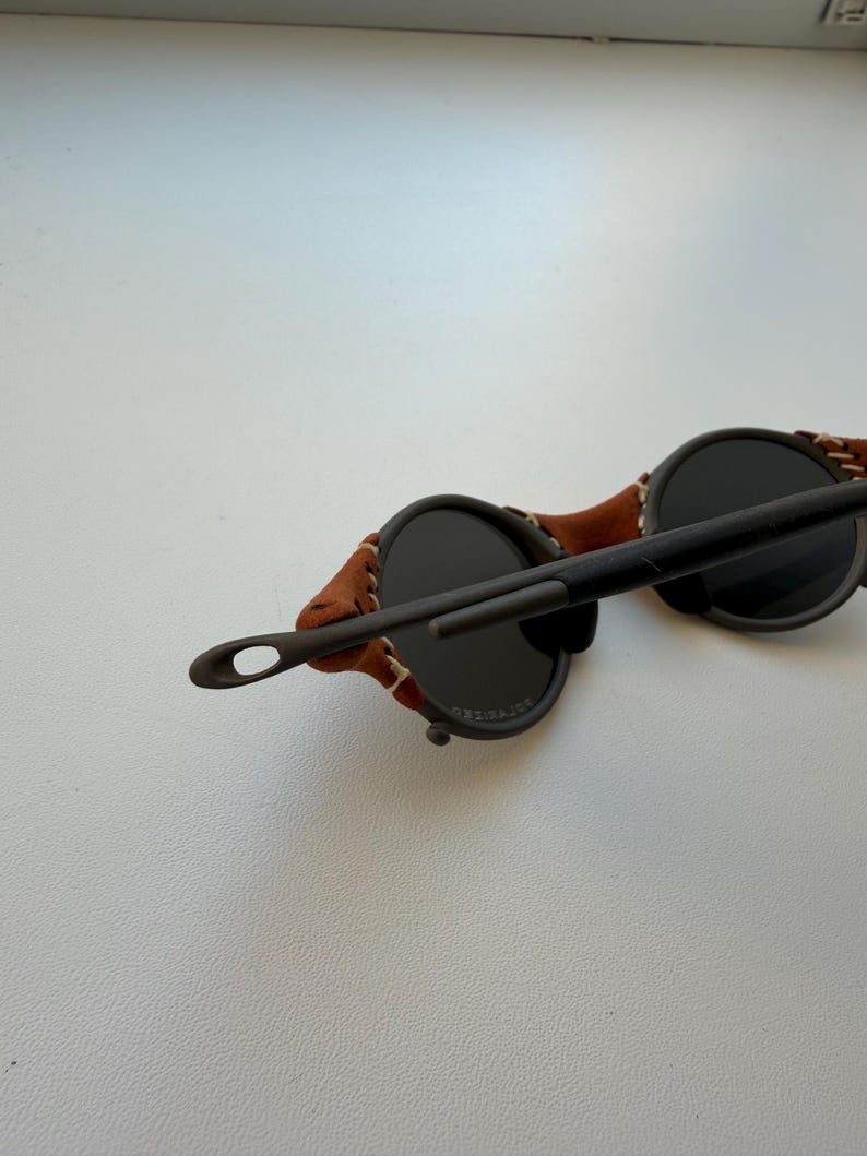 May include: Round sunglasses with dark lenses and a brown leather strap. The frame is dark, and the leather strap is stitched to the frame. The sunglasses have a unique, vintage style.