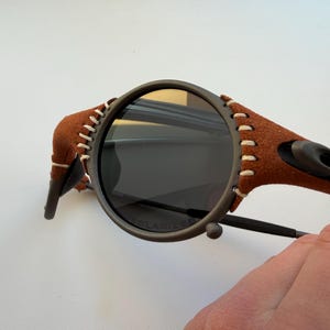 May include: Close-up of sunglasses with a dark gray frame and dark lenses. The temples are wrapped in brown suede with white stitching. The lenses are marked "POLARIZED".
