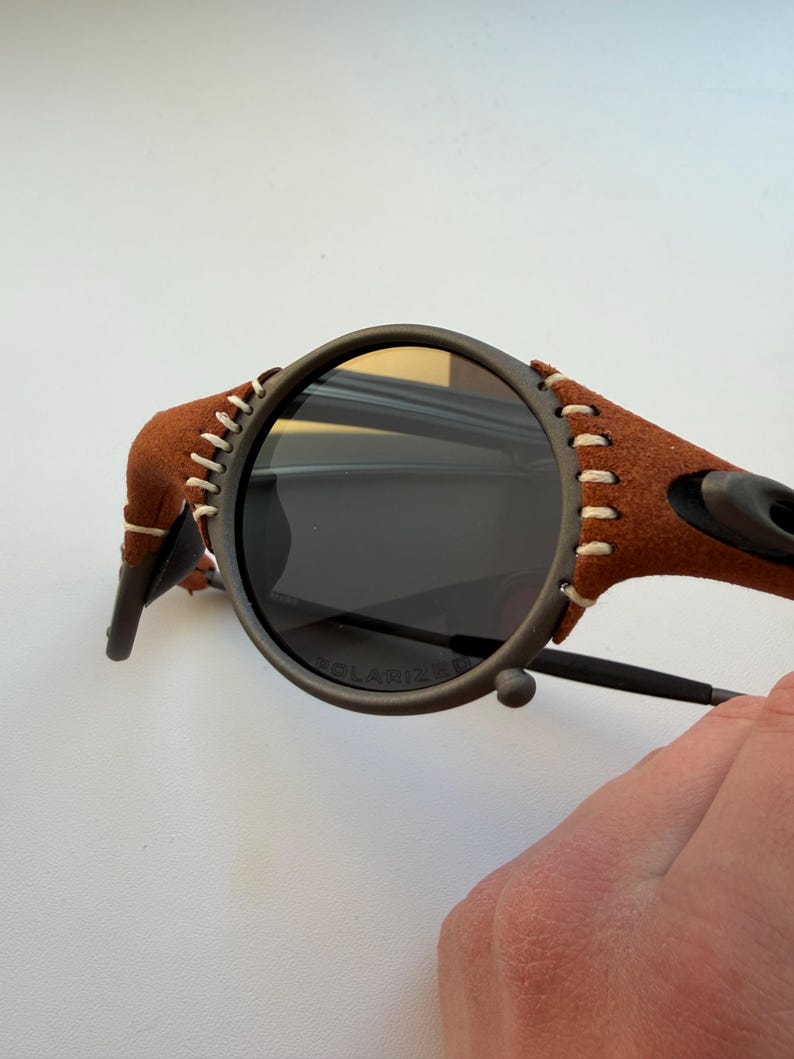 May include: Close-up of sunglasses with a dark gray frame and dark lenses. The temples are wrapped in brown suede with white stitching. The lenses are marked "POLARIZED".