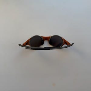 May include: Round sunglasses with dark lenses and a brown leather bridge. The leather is stitched to the frame. The arms are black and curve down. The sunglasses are set against a white background.