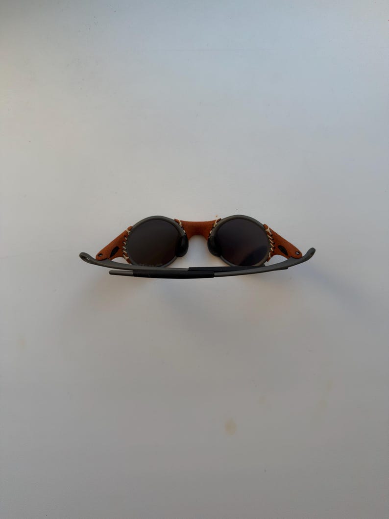 May include: Round sunglasses with dark lenses and a brown leather bridge. The leather is stitched to the frame. The arms are black and curve down. The sunglasses are set against a white background.