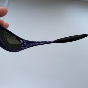 Puede incluir: Gafas de sol con montura morada y negra veteada. Las lentes son oscuras y las patillas tienen puntas de goma negra. Las gafas de sol est&aacute;n sujetas por una mano.