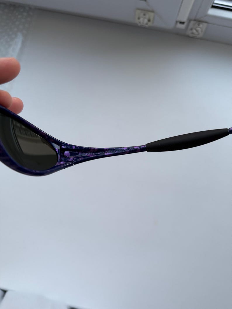 Puede incluir: Gafas de sol con montura morada y negra veteada. Las lentes son oscuras y las patillas tienen puntas de goma negra. Las gafas de sol est&aacute;n sujetas por una mano.