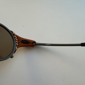 May include: Close-up of Oakley Mars sunglasses. The sunglasses have a dark grey frame with brown leather accents and stitching. The lenses are a dark tint. The word "MARS" is printed on the arm.