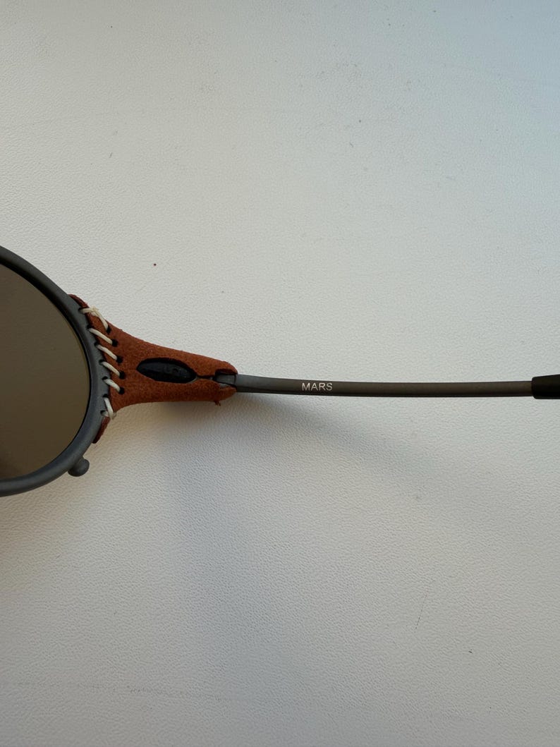 May include: Close-up of Oakley Mars sunglasses. The sunglasses have a dark grey frame with brown leather accents and stitching. The lenses are a dark tint. The word "MARS" is printed on the arm.
