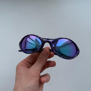 Puede incluir: Gafas de sol moradas y negras con lentes azules iridiscentes. Las gafas tienen un patr&oacute;n moteado en las monturas y un puente negro. Las lentes reflejan un degradado de tonos azules y morados. Las gafas son sostenidas por una mano.