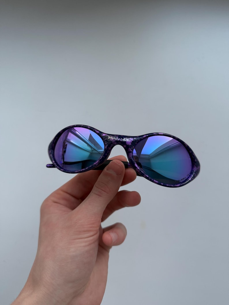 Puede incluir: Gafas de sol moradas y negras con lentes azules iridiscentes. Las gafas tienen un patr&oacute;n moteado en las monturas y un puente negro. Las lentes reflejan un degradado de tonos azules y morados. Las gafas son sostenidas por una mano.