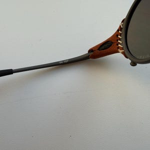 May include: Close-up of sunglasses with dark lenses and a brown leather accent stitched to the frame. The arm of the glasses is dark gray with the number "04-107" printed on it. The glasses are set against a light gray background.