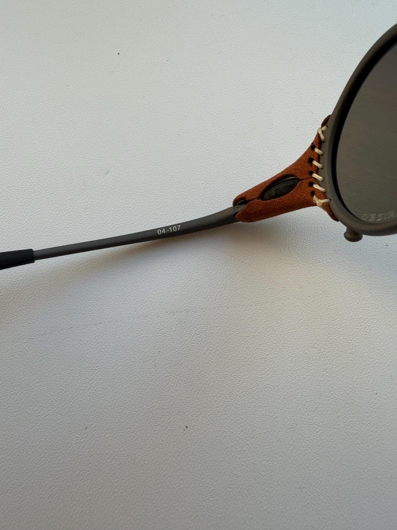 May include: Close-up of sunglasses with dark lenses and a brown leather accent stitched to the frame. The arm of the glasses is dark gray with the number "04-107" printed on it. The glasses are set against a light gray background.