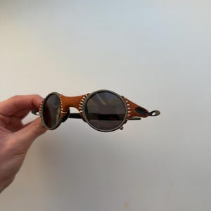 May include: Round sunglasses with dark lenses and a brown leather and metal frame. The frame features visible stitching and a unique design. The sunglasses have a steampunk aesthetic.