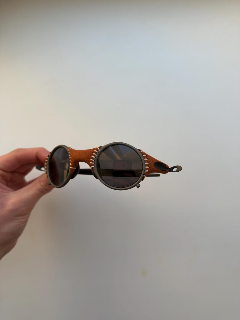 May include: Round sunglasses with dark lenses and a brown leather and metal frame. The frame features visible stitching and a unique design. The sunglasses have a steampunk aesthetic.