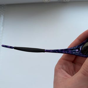 Puede incluir: Primer plano de gafas de sol con montura moteada morada y negra. El brazo de las gafas de sol est&aacute; sujeto por una mano. El brazo tiene un agarre de goma negro cerca de la oreja.