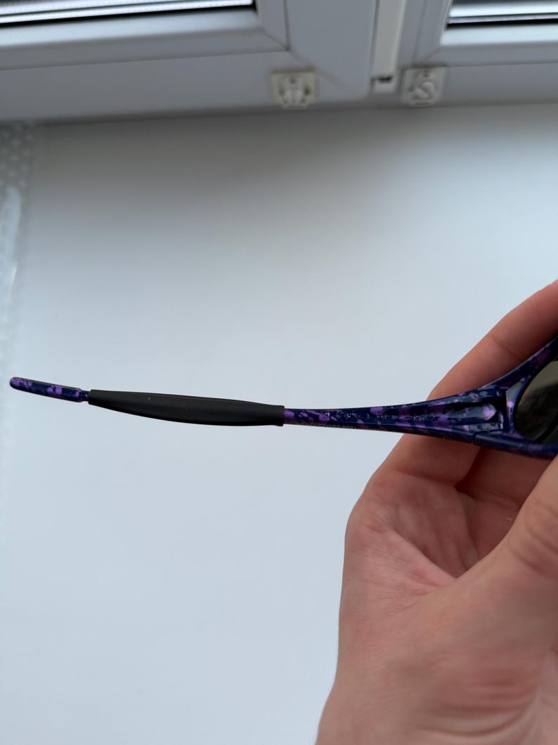 Puede incluir: Primer plano de gafas de sol con montura moteada morada y negra. El brazo de las gafas de sol est&aacute; sujeto por una mano. El brazo tiene un agarre de goma negro cerca de la oreja.