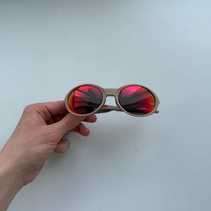 Op de afbeelding: Zonnebril met een beige montuur en ronde lenzen die rode en oranje kleuren reflecteren. Het ontwerp is klassiek. De afbeelding is genomen tegen een effen witte achtergrond. Geschikt voor verschillende stijlen.