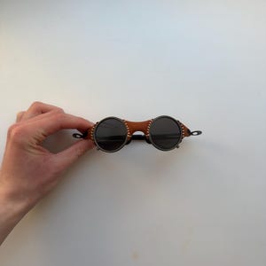 May include: Round sunglasses with dark lenses and a brown leather bridge. The frames are a dark metal color, and the leather bridge is stitched with white thread. The sunglasses have a unique, vintage style.