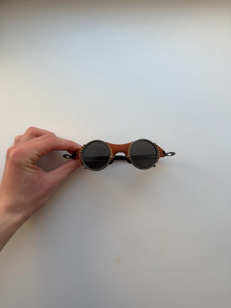 May include: Round sunglasses with dark lenses and a brown leather bridge. The frames are a dark metal color, and the leather bridge is stitched with white thread. The sunglasses have a unique, vintage style.