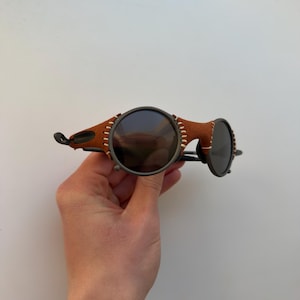May include: Round sunglasses with dark lenses and a brown leather bridge. The frame is a dark metal with visible stitching on the leather. The sunglasses are held by a hand.