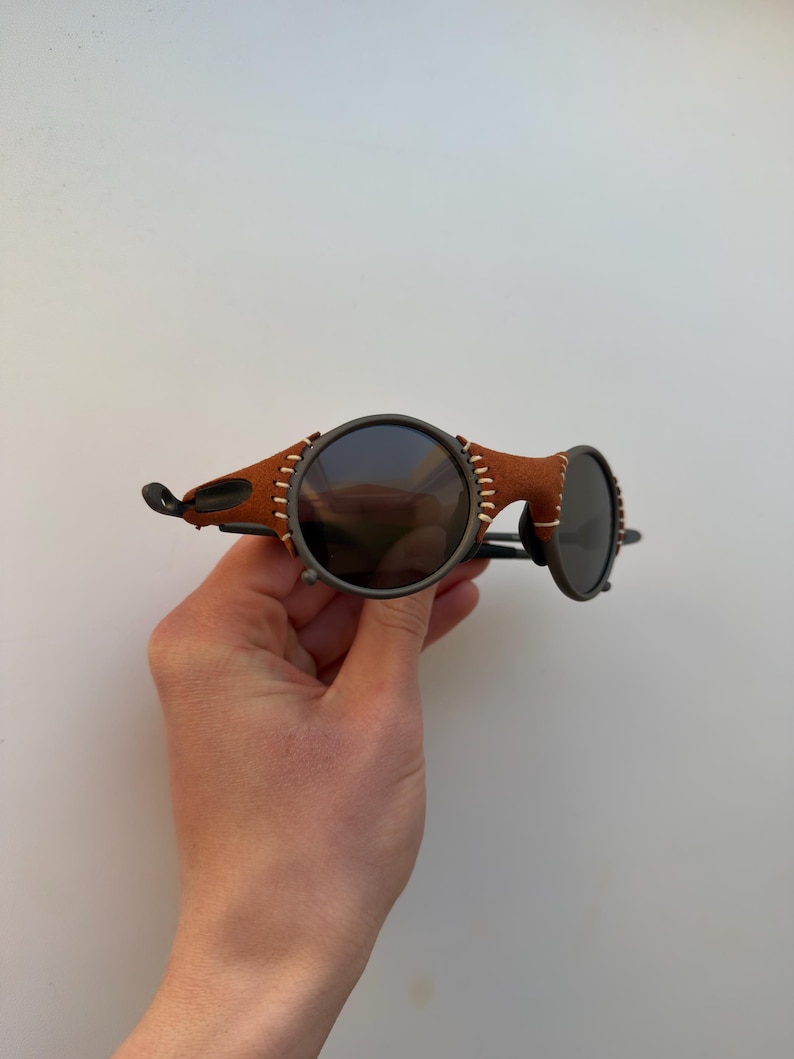 May include: Round sunglasses with dark lenses and a brown leather bridge. The frame is a dark metal with visible stitching on the leather. The sunglasses are held by a hand.