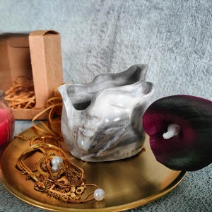 Może przedstawiać: Dekoracyjna aranżacja z wazonem z efektem marmuru, złotą tacą z biżuterią, czerwoną świecą w szklanym słoiku, brązowym pudełkiem prezentowym i ciemnofioletową kalą. Przedmioty ułożone są na szarej powierzchni.