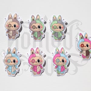 May include: A collection of eight whimsical sticker designs featuring stitched bunny characters in various color combinations. Each bunny has button eyes, a stitched mouth, and is adorned with pins, creating a unique, handcrafted aesthetic.