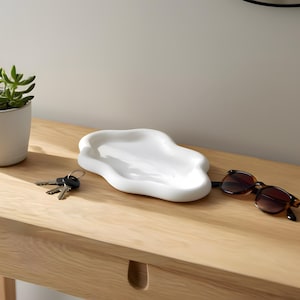 May include: A white, organically shaped ceramic tray, sunglasses, keys, and a small potted succulent plant sit on a light wood surface. The sunglasses have brown frames and lenses. The plant is in a white pot.