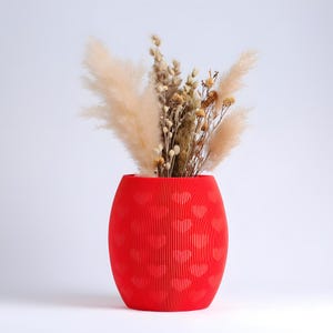 May include: A vibrant red vase with a textured, ribbed design and heart-shaped cutouts. The vase is filled with dried beige and brown ornamental grasses and flowers, creating a decorative floral arrangement. The vase is a decorative home accent.