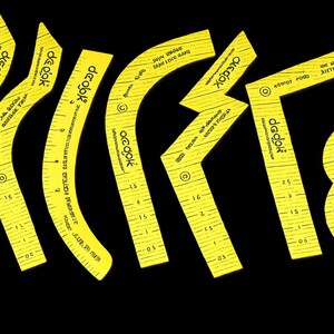 Puede incluir: Patrones de costura de plástico amarillo con bordes curvos y angulados, marcados con medidas en centímetros y el nombre de la marca "deok". Las piezas están dispuestas sobre un fondo negro.