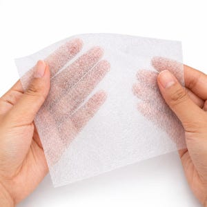 Puede incluir: Una hoja rectangular delgada de tela no tejida blanca semitransparente con una textura fibrosa similar a una malla. El material es ligero y poroso, lo que permite una visibilidad parcial a través de la superficie de la hoja.