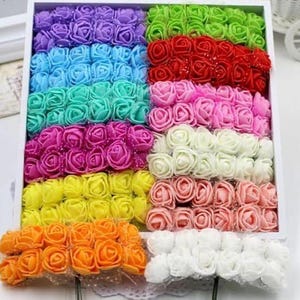 May include: A white box filled with colorful artificial rose buds. The roses are arranged in rows and include shades of purple, blue, green, red, pink, yellow, orange, and white. These decorative flowers are ideal for crafts and DIY projects.