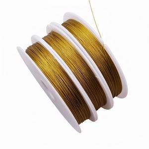 May include: Four spools of gold-colored beading wire are displayed against a white background. The wire is tightly wound on white plastic spools. A single strand of wire extends from one of the spools.