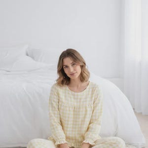 May include: A light yellow gingham pajama set. The long-sleeved top has a round neckline and ruffled cuffs. The matching pants are also gingham. The person is sitting on a white rug in front of a bed.
