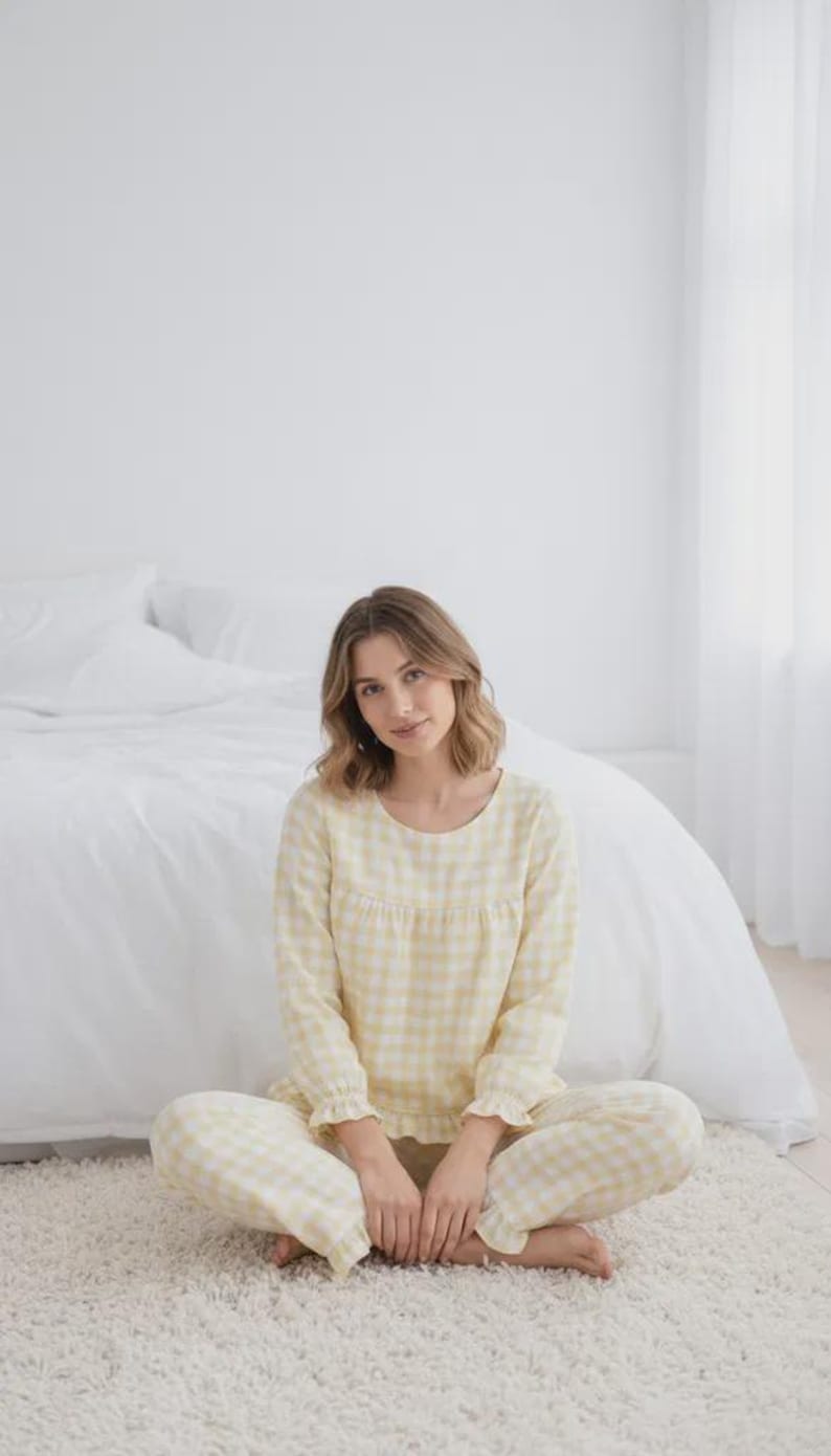 May include: A light yellow gingham pajama set. The long-sleeved top has a round neckline and ruffled cuffs. The matching pants are also gingham. The person is sitting on a white rug in front of a bed.