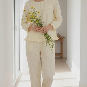 May include: Cream-colored striped pajama set. The long-sleeved top and pants have a ruffled trim. The person in the image is holding a bouquet of yellow flowers. The pajamas are made of a soft, lightweight fabric, perfect for lounging or sleeping.