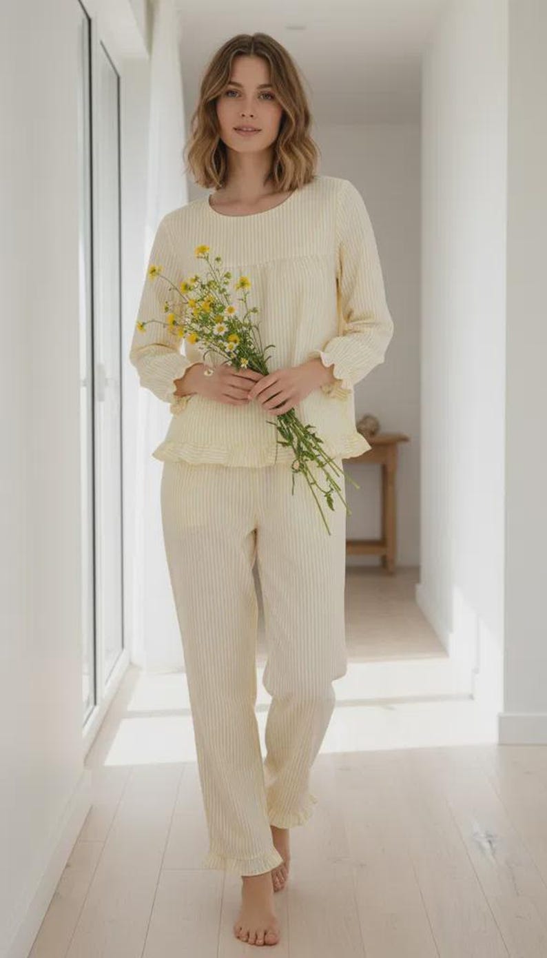 May include: Cream-colored striped pajama set. The long-sleeved top and pants have a ruffled trim. The person in the image is holding a bouquet of yellow flowers. The pajamas are made of a soft, lightweight fabric, perfect for lounging or sleeping.
