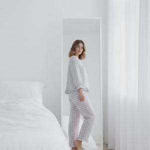 May include: A person wearing a light gray and pink gingham pajama set, consisting of a long-sleeved top and pants. The pajamas have a ruffled hem and are reflected in a full-length mirror. The room has white walls, a bed with white linens, and sheer curtains.