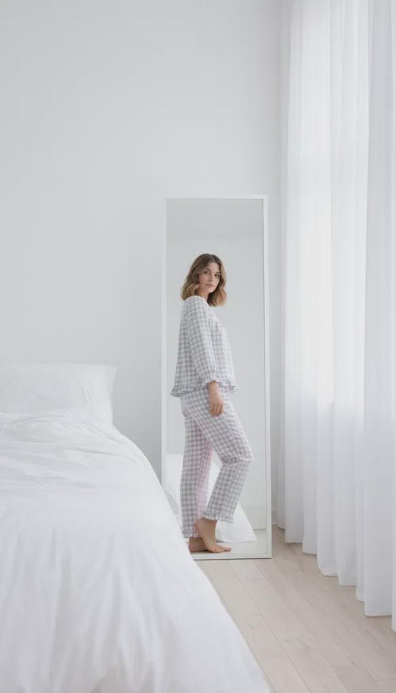 May include: A person wearing a light gray and pink gingham pajama set, consisting of a long-sleeved top and pants. The pajamas have a ruffled hem and are reflected in a full-length mirror. The room has white walls, a bed with white linens, and sheer curtains.