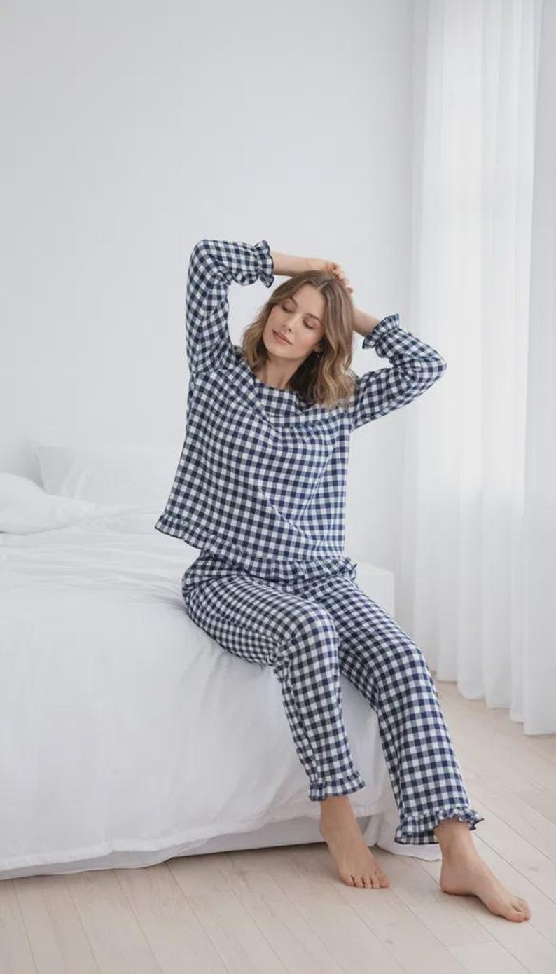 May include: A two-piece pajama set in a navy and white gingham pattern. The long-sleeved top and pants have a relaxed fit. The pajamas have ruffled trim at the cuffs and ankles. The person is sitting on a white bed.