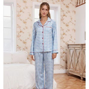 Op de afbeelding: Een lichtblauw en wit geruite pyjamaset. De top met lange mouwen heeft rode biezen rond de kraag en manchetten, met rode knopen. De bijpassende broek heeft een vergelijkbaar ruitpatroon en rode biezen. De pyjama's zijn gemaakt van zacht, comfortabel materiaal.