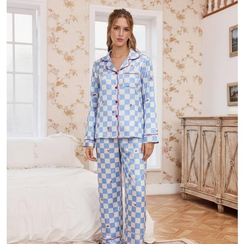 Op de afbeelding: Een lichtblauw en wit geruit pyjamaset. De button-down shirt met lange mouwen en bijpassende broek hebben rode biezen en kleine rode hartdetails. De pyjama is gemaakt van een zijdeachtig materiaal.