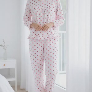 May include: A two-piece pajama set in white with a repeating pattern of small red hearts. The long-sleeved top has a ruffled hem and the pants have a matching ruffle at the ankles. The pajamas are made of a soft, lightweight fabric.