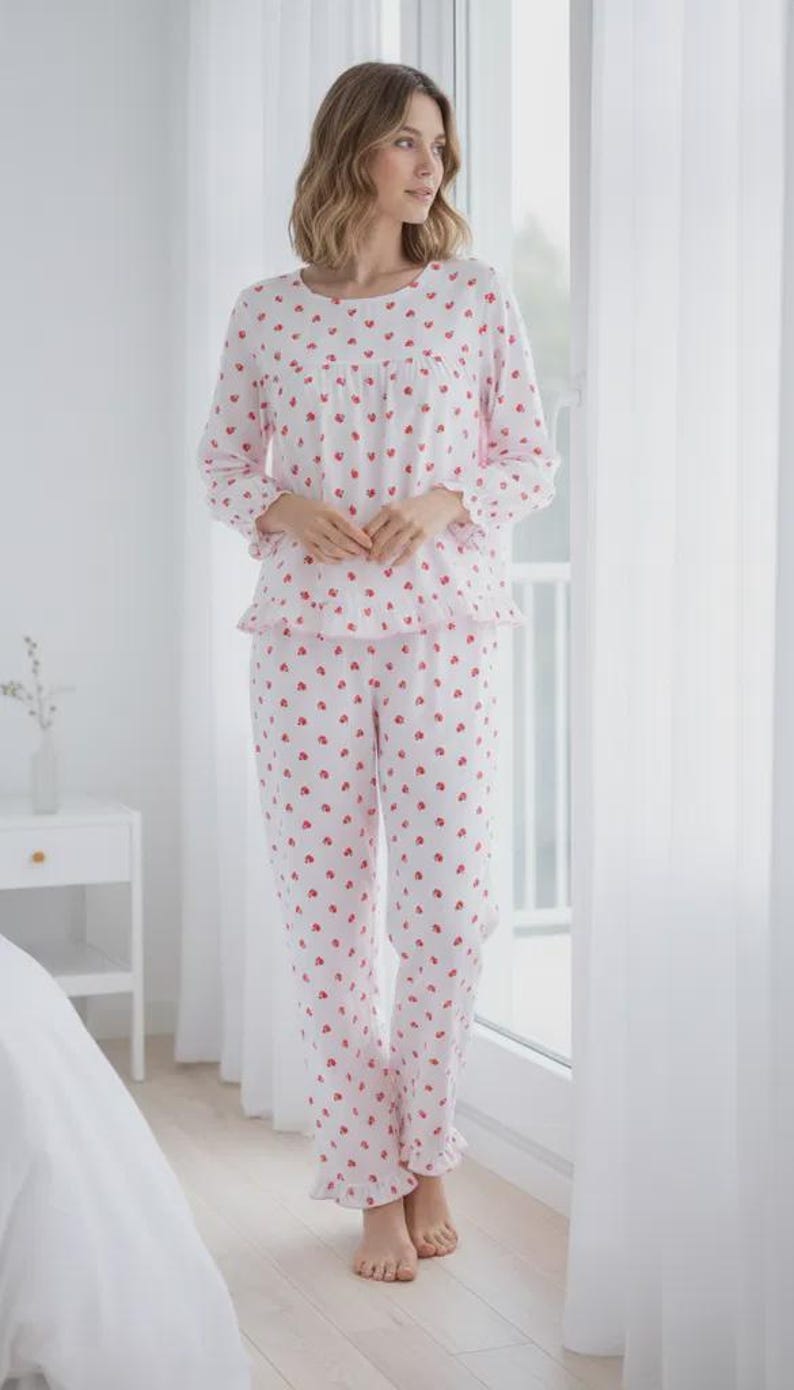 May include: A two-piece pajama set in white with a repeating pattern of small red hearts. The long-sleeved top has a ruffled hem and the pants have a matching ruffle at the ankles. The pajamas are made of a soft, lightweight fabric.